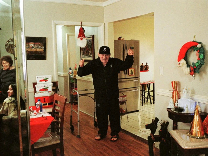 Man in a black tracksuit and baseball cap stands with raised arms indoors, celebrating near Christmas decorations with family members.
