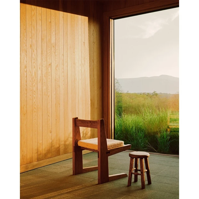 Interior room featuring a wooden chair and stool near a large window, showcasing an outdoor landscape view with greenery and distant mountains.