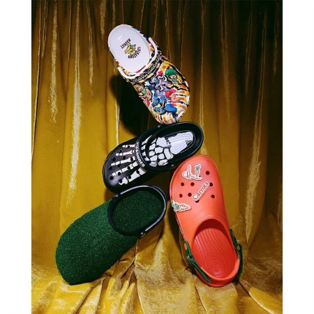 Four Crocs clogs in a vertical composition against a golden, shimmering curtain backdrop. Each clog has a unique design.