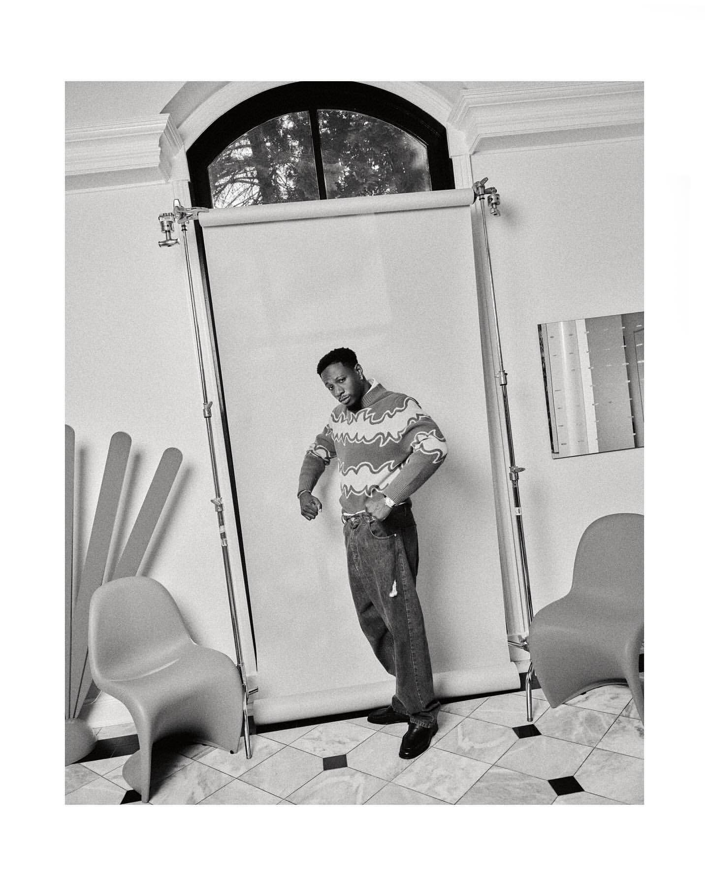 Black and white portrait of a Black man in patterned sweater and jeans standing in a photo studio with chairs and backdrop.
