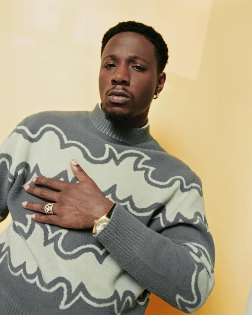 Adult man poses in a grey and light green patterned sweater against a pale yellow studio backdrop, showcasing a ring and watch.