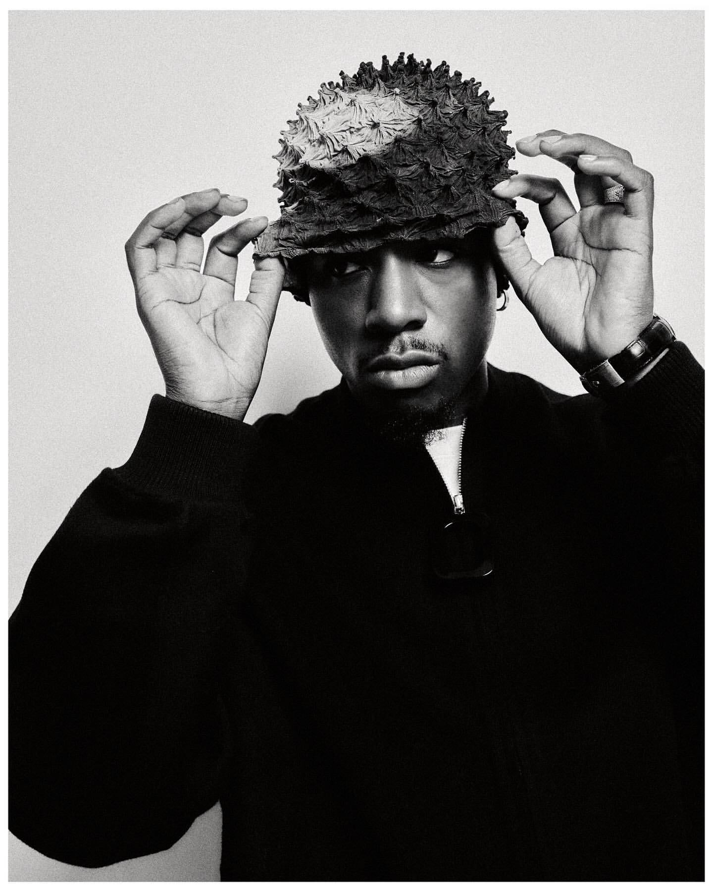 Monochromatic portrait of a black male wearing a textured bucket hat and dark sweater, looking directly at the camera in a studio setting.