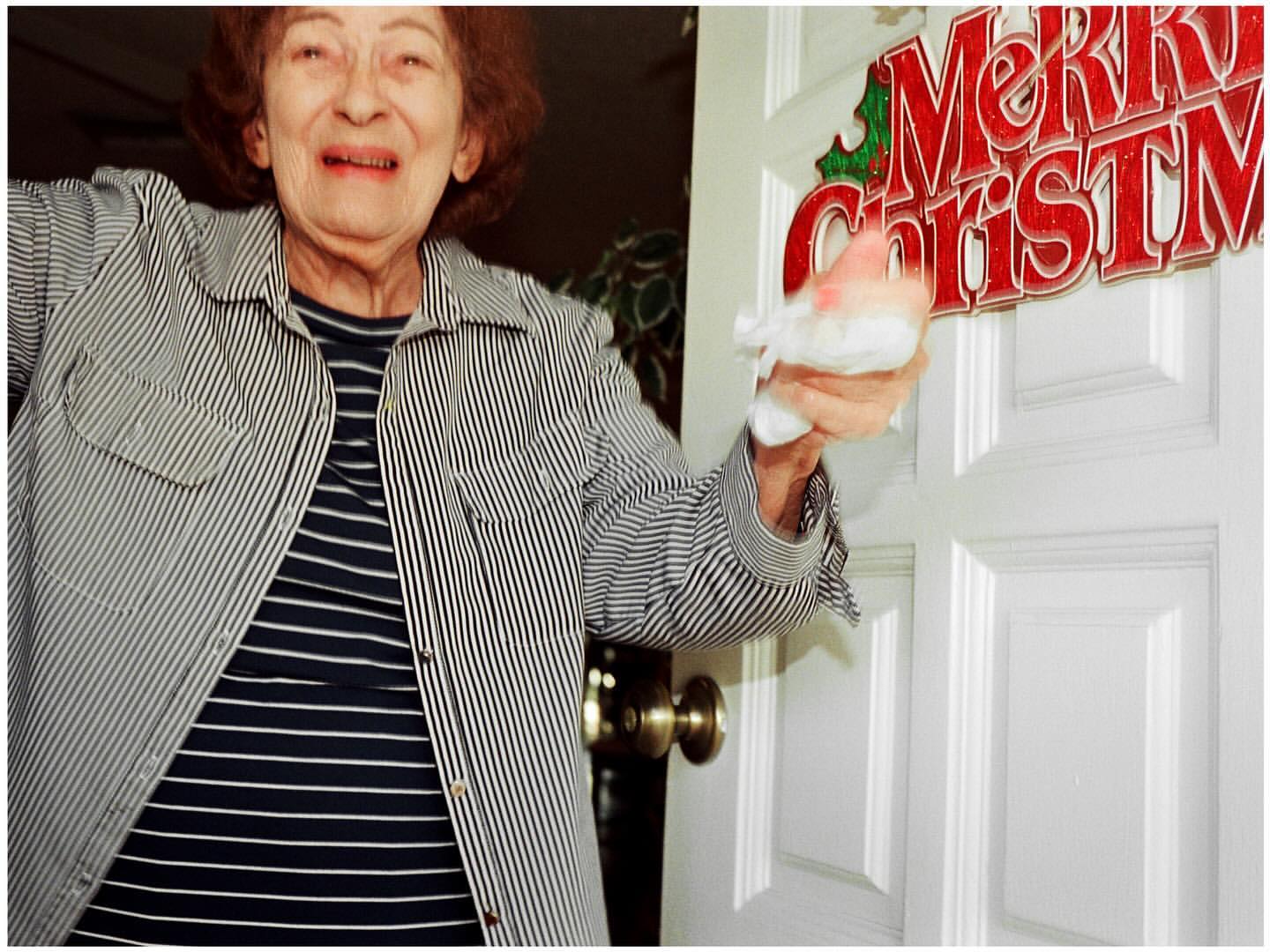 Elderly woman in a striped shirt smiles and holds a Santa Claus figurine by a white door decorated with a 'Merry Christmas' sign indoors.