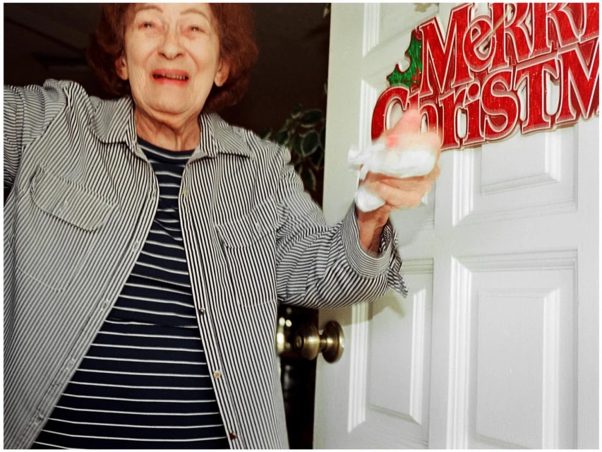 Elderly woman in a striped shirt smiles and holds a Santa Claus figurine by a white door decorated with a 'Merry Christmas' sign indoors.