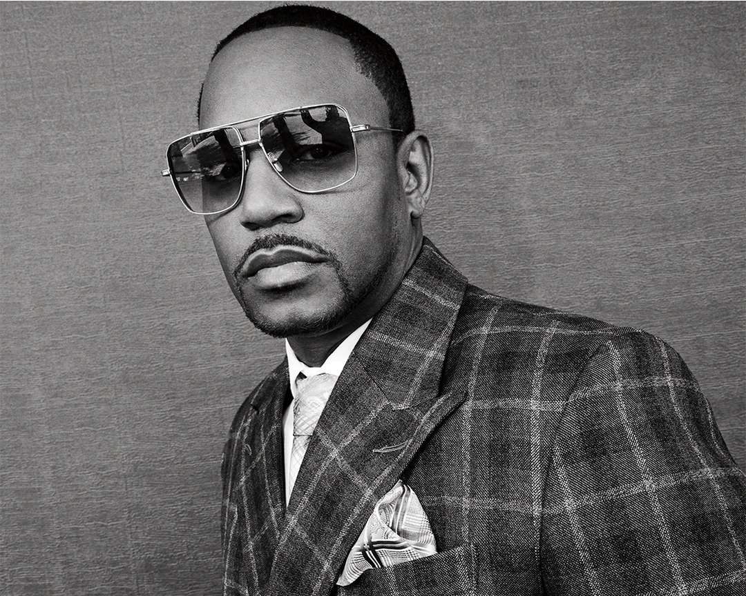 Portrait of an adult Black man in a grey plaid suit with sunglasses, a tie, and a handkerchief, against a grey background.