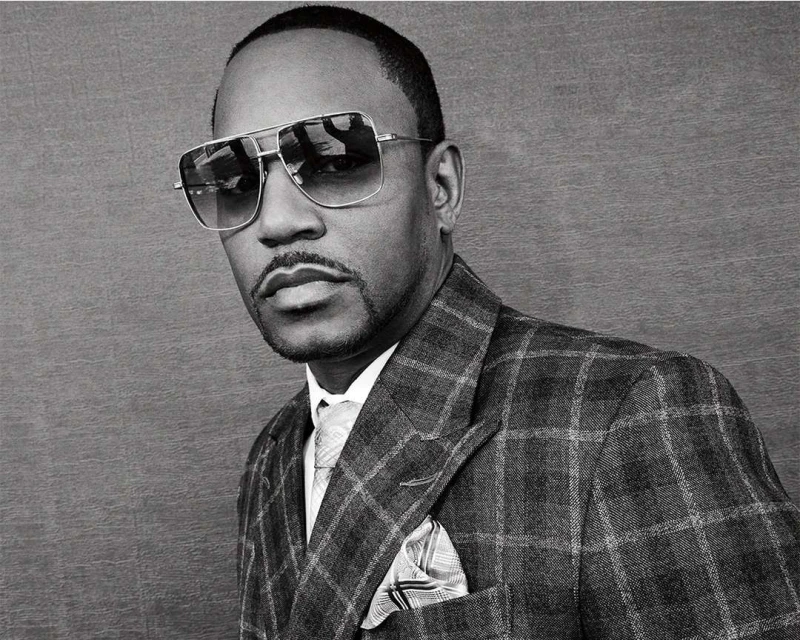 Portrait of an adult Black man in a grey plaid suit with sunglasses, a tie, and a handkerchief, against a grey background.