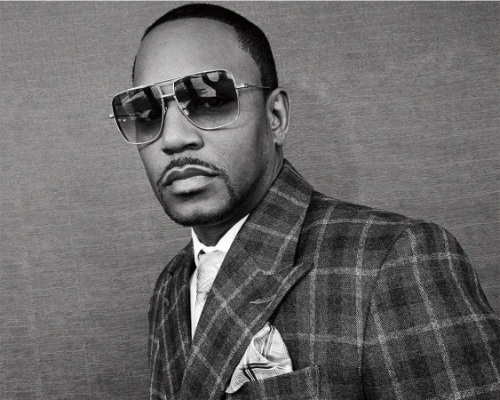 Portrait of an adult Black man in a grey plaid suit with sunglasses, a tie, and a handkerchief, against a grey background.