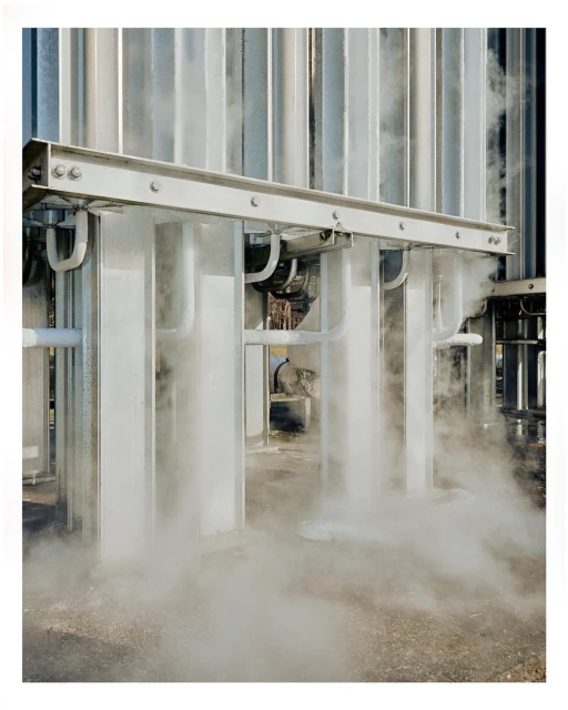 Industrial scene featuring a network of metal pipes with visible vapor or condensation, suggesting a cooling or refrigeration process.