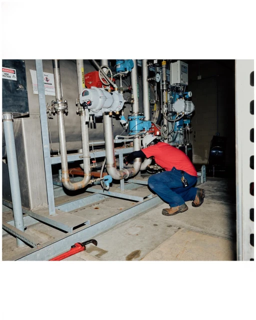 Man in red shirt crouching by industrial pipes, wearing gloves and jeans, working on plumbing with silver tanks in the background.