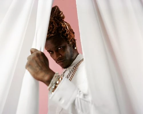 Portrait of a stylish young adult male peeking from behind white curtains, wearing a white button-down shirt and gold jewellery against a pink backdrop.