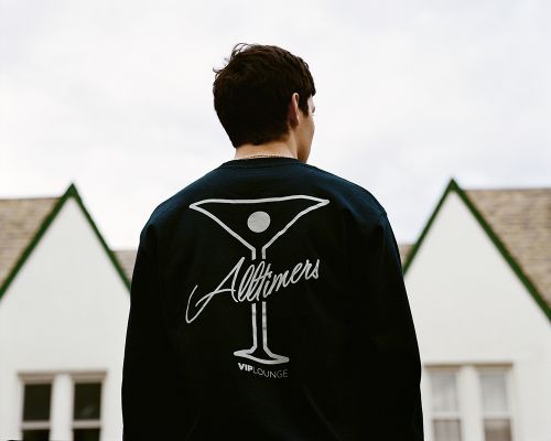 Back view of a young adult wearing a black shirt with 'Alltimers VIP Lounge' design, standing outdoors with a white building behind him.