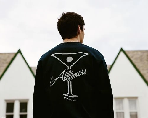 Back view of a young adult wearing a black shirt with 'Alltimers VIP Lounge' design, standing outdoors with a white building behind him.