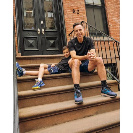 Adult male and child sit on brown stone steps outside a red brick building, wearing athleisure wear and athletic shoes in daylight.