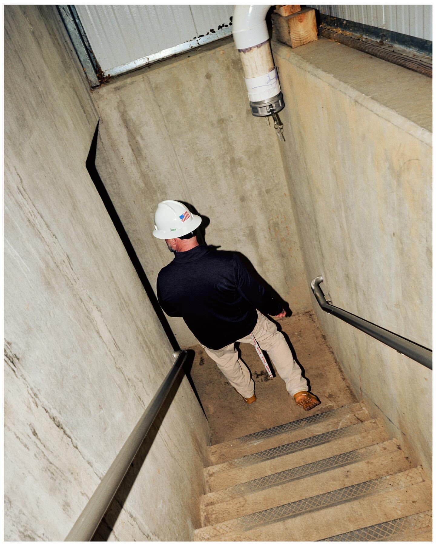 Man with hardhat descends metal stairs in concrete industrial setting, illuminated by overhead lighting, holding onto a metal handrail.