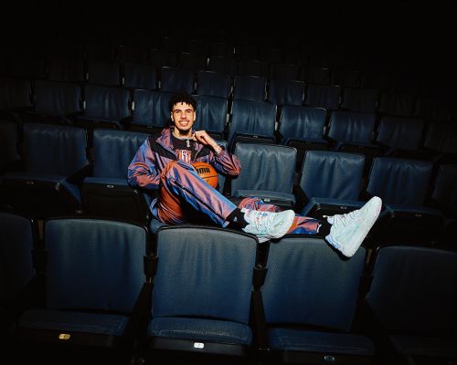 Young man in iridescent tracksuit and sneakers lounges in empty theater seats, holding basketball, bathed in soft light.