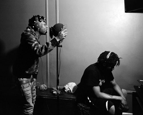 Monochrome portrait of two adult male musicians in a recording studio, one singing into a microphone and the other seated nearby.