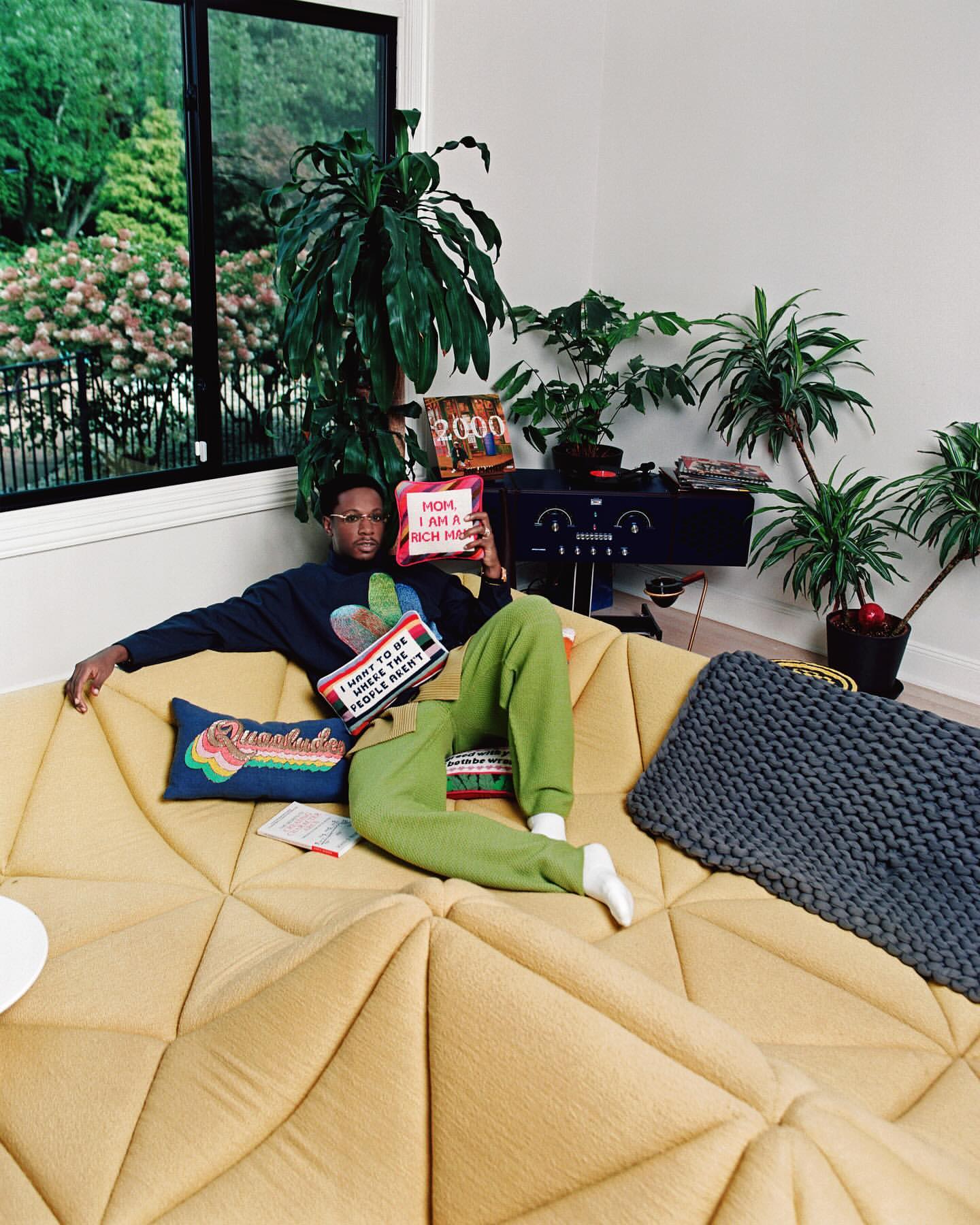 Man relaxes on a modern yellow couch in a room filled with houseplants, holding decorative pillows, creating a cozy, indoor scene.