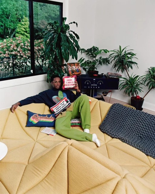 Man relaxes on a modern yellow couch in a room filled with houseplants, holding decorative pillows, creating a cozy, indoor scene.