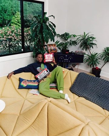 Man relaxes on a modern yellow couch in a room filled with houseplants, holding decorative pillows, creating a cozy, indoor scene.