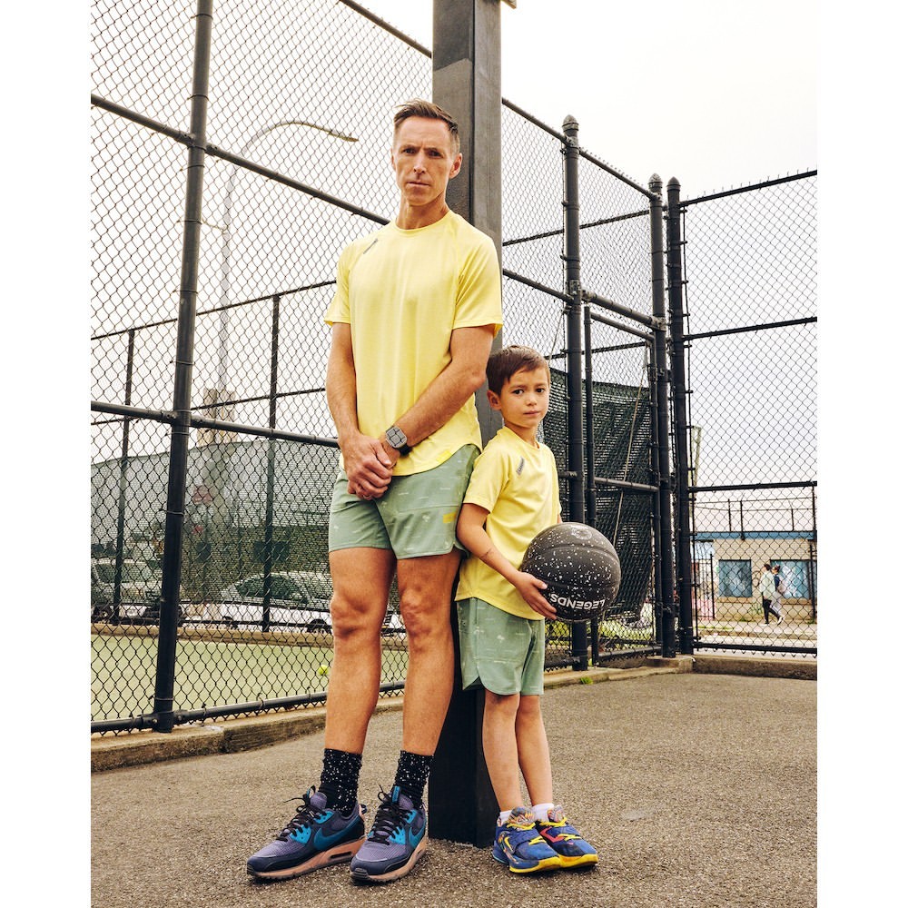 An adult and a child stand outdoors on a basketball court with a chain-link fence in the background. The child is holding a basketball.
