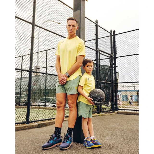 An adult and a child stand outdoors on a basketball court with a chain-link fence in the background. The child is holding a basketball.