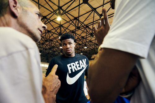 Low angle of basketball player Giannis Antetokounmpo in a black t-shirt surrounded by two people inside a gymnasium.