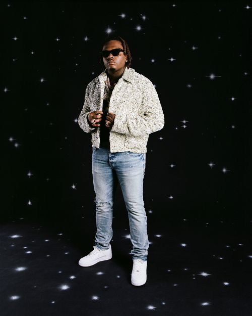 Full-length portrait of a young adult male wearing sunglasses, a tweed jacket, blue jeans, and white sneakers standing against a starscape background.