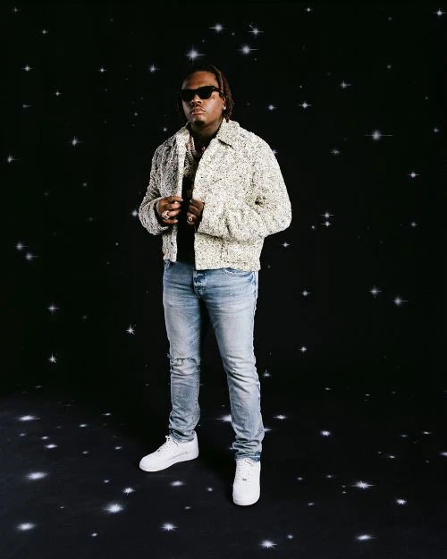 Full-length portrait of a young adult male wearing sunglasses, a tweed jacket, blue jeans, and white sneakers standing against a starscape background.