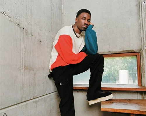 Man posing indoors in casual, color-blocked sweater and black pants, leaning against concrete wall with window on wooden steps.