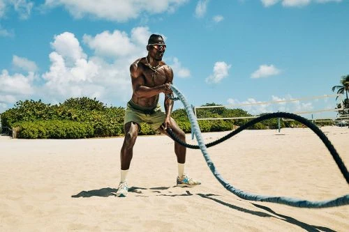 Shirtless athletic man working out with battle ropes on beach in bright sunlight against sky with scattered clouds.