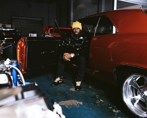 Man in yellow beanie and black puffer jacket seated in a red vintage car with the door open, inside of auto repair shop.