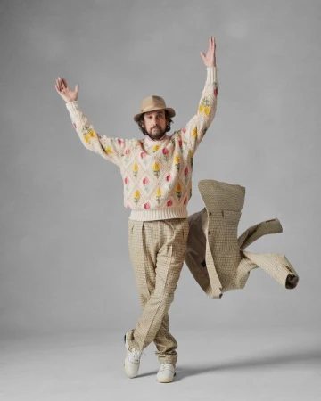 Man wearing a patterned sweater, trousers, fedora, and holding a jacket in the air against a grey studio backdrop