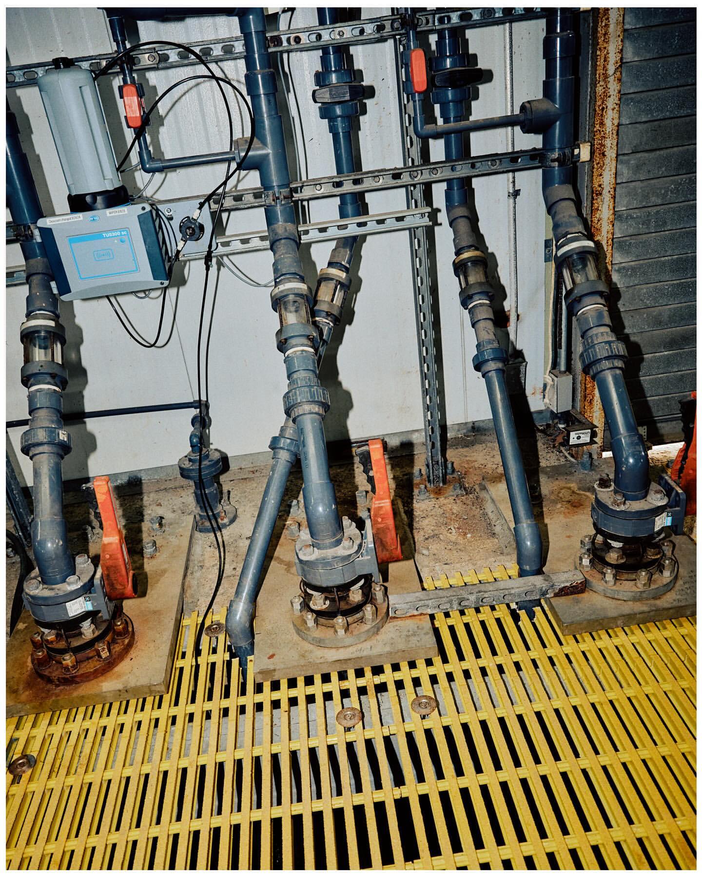 Industrial pipes and valves connect to machinery on a yellow platform with a white wall in the background.