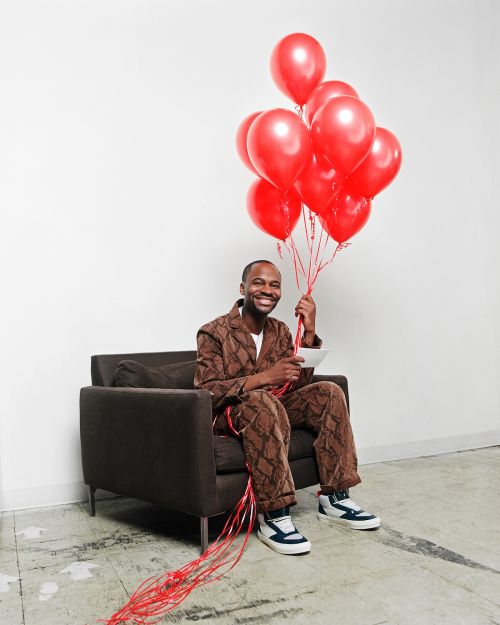 Smiling man sits holding a bunch of red balloons in a studio, wearing a brown patterned jacket and pants with white sneakers.