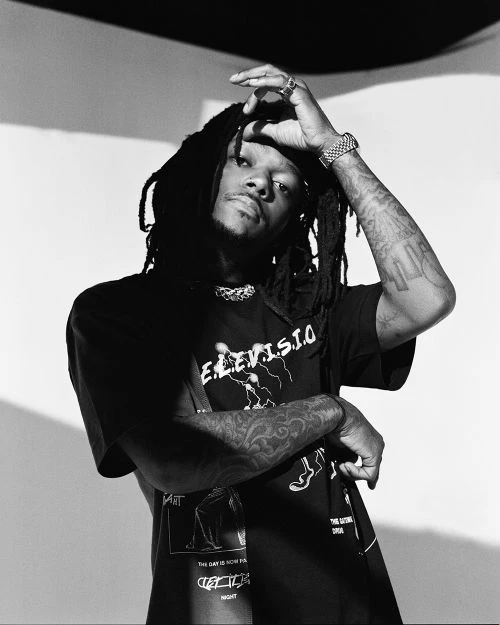 Monochrome portrait of Black male model with dreadlocks, graphic tee, and tattoos posing in a studio setting with hand on head.