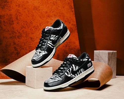 Two black and white Nike Dunk Low zebra print sneakers are displayed on a light tan platform with an orange textured background.
