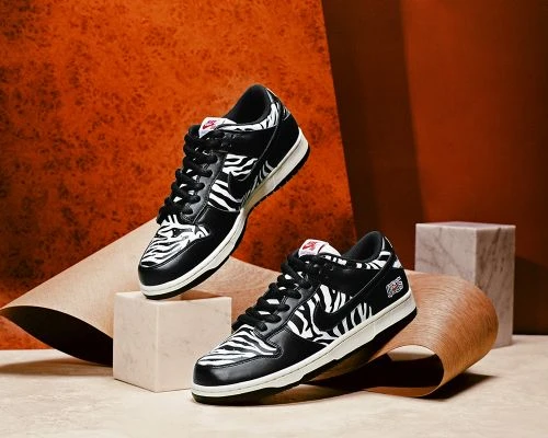 Two black and white Nike Dunk Low zebra print sneakers are displayed on a light tan platform with an orange textured background.