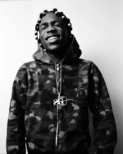 Black and white portrait of a smiling African American man wearing a camouflage hooded jacket and a pendant necklace.