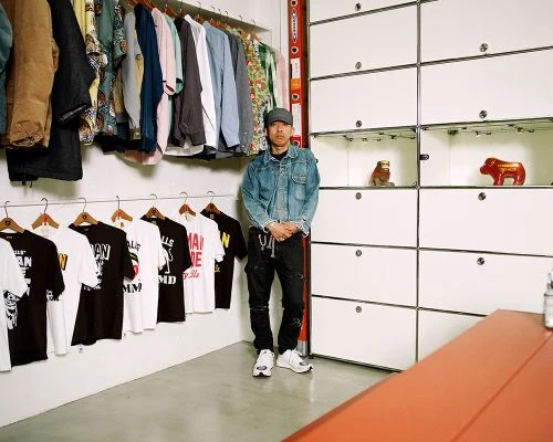 Man in denim jacket stands in a retail space surrounded by hanging clothes, t-shirts and a modular display cabinet with decorative items.