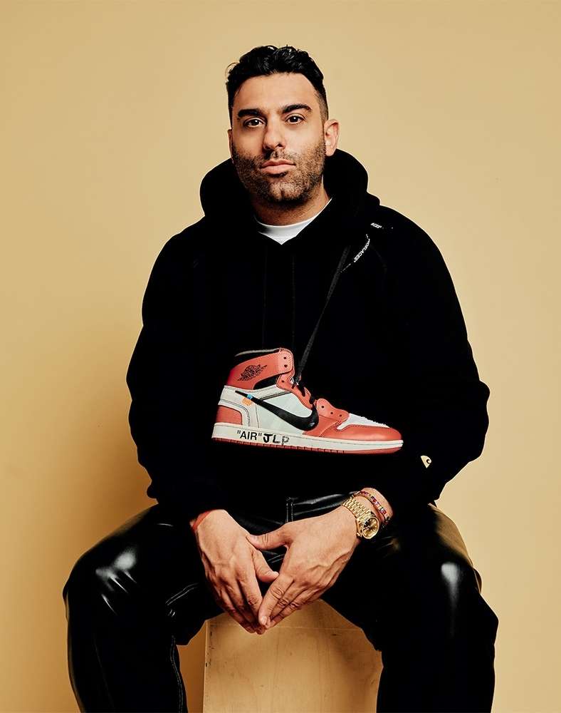 Adult male in black hoodie posing with high-top sneaker purse in a studio with tan background. Portrait shot, shiny black pants.