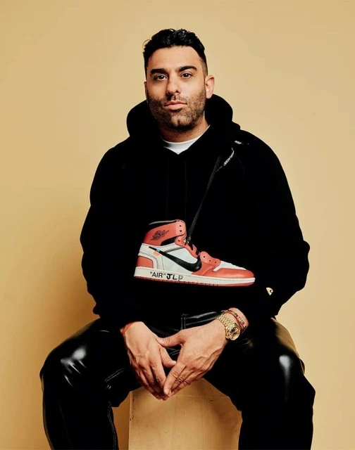 Adult male in black hoodie posing with high-top sneaker purse in a studio with tan background. Portrait shot, shiny black pants.