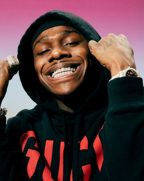 Portrait of DaBaby smiling broadly, wearing a black hooded sweatshirt with a prominent red Gucci logo, accessorized with diamond jewelry.