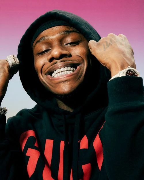 Portrait of DaBaby smiling broadly, wearing a black hooded sweatshirt with a prominent red Gucci logo, accessorized with diamond jewelry.