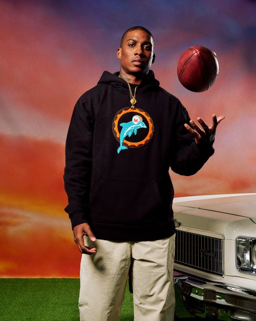 Adult male in a black hoodie with a dolphin graphic, holding a football in the air in front of a car and sky background.