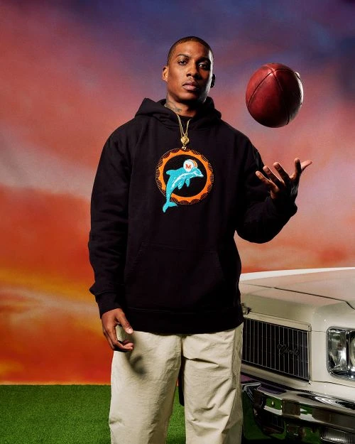 Adult male in a black hoodie with a dolphin graphic, holding a football in the air in front of a car and sky background.