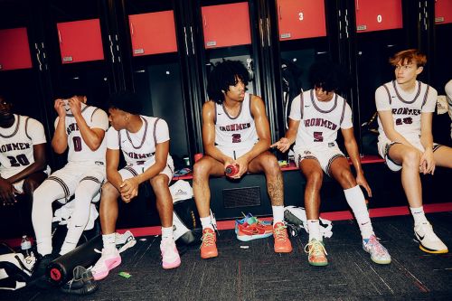 Five male basketball players in uniform sit on a bench in their locker room, resting and talking before or after a game.