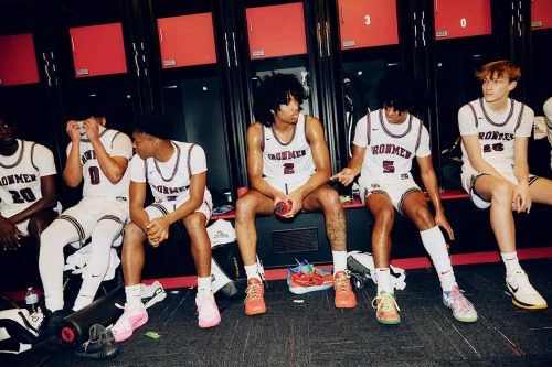 Five male basketball players in uniform sit on a bench in their locker room, resting and talking before or after a game.