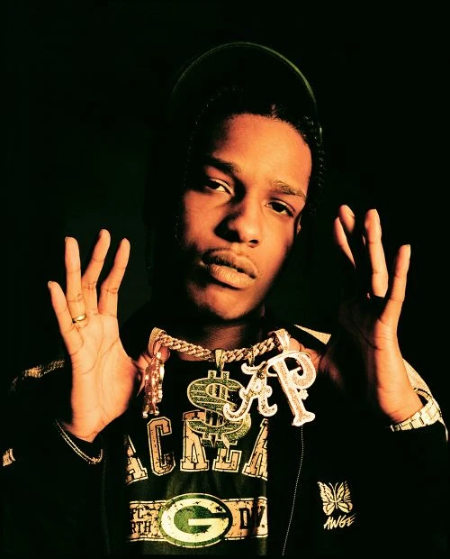 Portrait of an African American man wearing a black jacket and gold chains, holding up both hands against a dark backdrop.