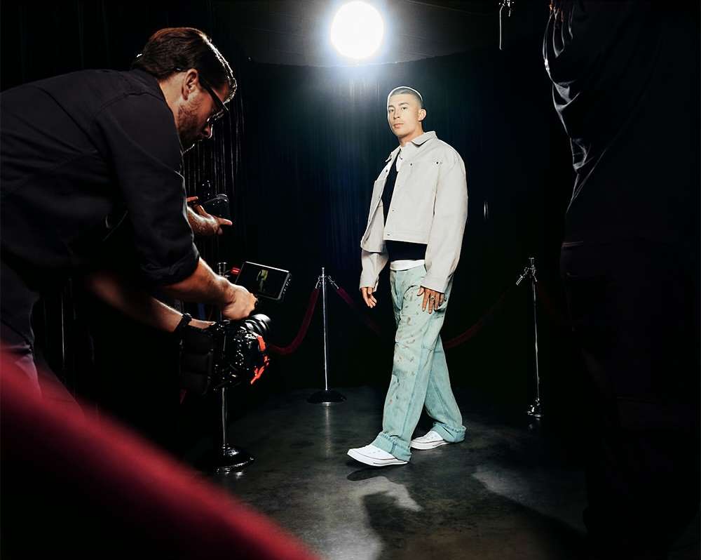 Man in a jacket walking towards the camera, being filmed by a cameraman, in a studio with a spotlight and red ropes.