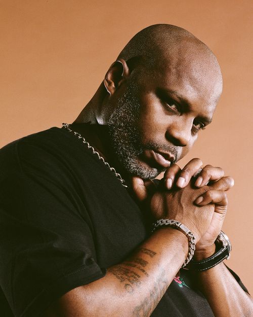 Portrait of African-American man, DMX, with hands together near face, wearing a black t-shirt, chain necklace, and bracelet against tan background.